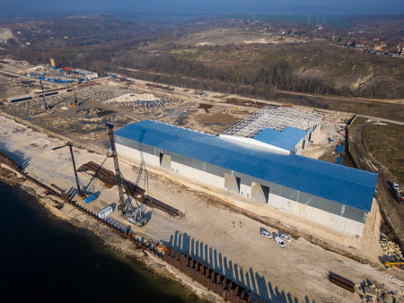 A construction site with a partially built warehouse featuring a steel framework and metal roof. Workers and heavy machinery operate on-site, showcasing industrial progress and structural developmentの写真素材