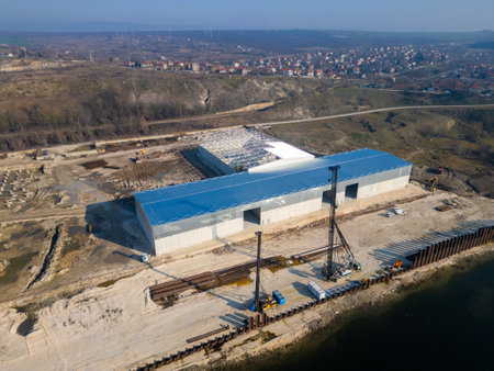A construction site with a partially built warehouse featuring a steel framework and metal roof. Workers and heavy machinery operate on-site, showcasing industrial progress and structural developmentの写真素材