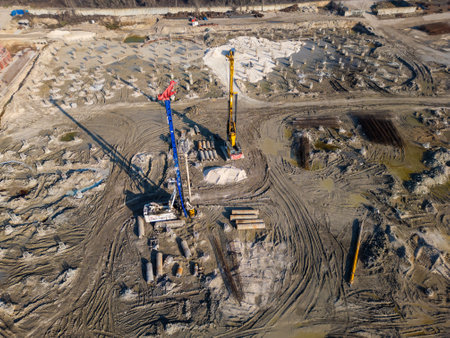 A construction site with heavy machinery drilling and preparing foundations. Piles, rebar, and muddy tracks cover the ground, showcasing progress in the industrial foundation development processの写真素材