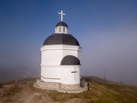 A small white church in Bulgaria stands on a hill, surrounded by fog and an expansive landscape. Its black domes and cross create a serene, spiritual atmosphere against the blue sky and misty valleyの写真素材