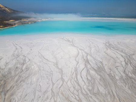 A turquoise tailings pond near a cement factory, surrounded by rugged hills. The vivid water contrasts with the barren land, creating a striking yet industrial scene of environmental impactの写真素材