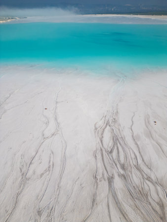 A turquoise tailings pond near a cement factory, surrounded by rugged hills. The vivid water contrasts with the barren land, creating a striking yet industrial scene of environmental impactの写真素材