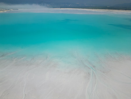 A turquoise tailings pond near a cement factory, surrounded by rugged hills. The vivid water contrasts with the barren land, creating a striking yet industrial scene of environmental impactの写真素材