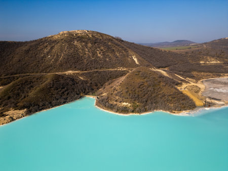 A turquoise tailings pond near a cement factory, surrounded by rugged hills. The vivid water contrasts with the barren land, creating a striking yet industrial scene of environmental impactの写真素材