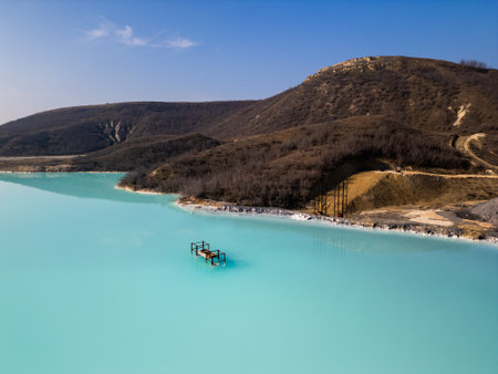 A turquoise pond near a cement factory, surrounded by rugged hills. The vivid water contrasts with the barren land, creating a striking yet industrial sceneの写真素材