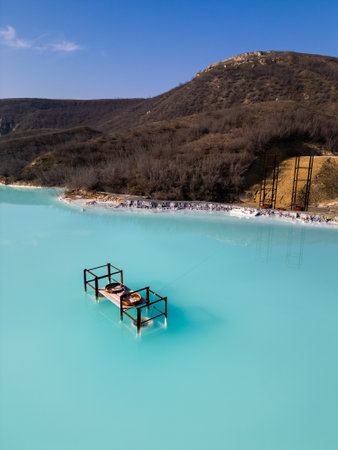 A turquoise tailings pond near a cement factory, surrounded by rugged hills. The vivid water contrasts with the barren land, creating a striking yet industrial scene of environmental impactの写真素材