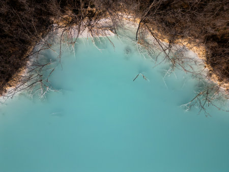 A turquoise tailings pond near a cement factory, surrounded by rugged hills. The vivid water contrasts with the barren land, creating a striking yet industrial scene of environmental impactの写真素材