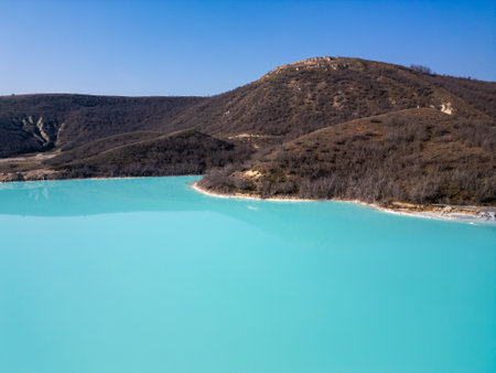 A turquoise tailings pond near a cement factory, surrounded by rugged hills. The vivid water contrasts with the barren land, creating a striking yet industrial scene of environmental impactの写真素材