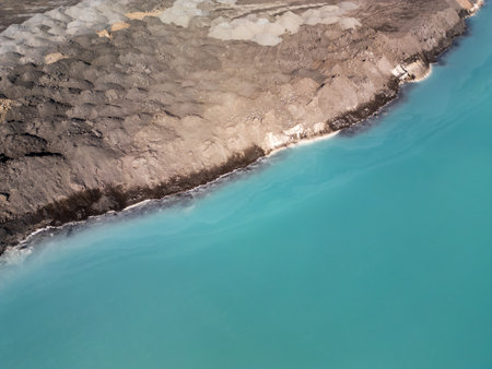 A turquoise tailings pond near a cement factory, surrounded by rugged hills. The vivid water contrasts with the barren land, creating a striking yet industrial scene of environmental impactの写真素材
