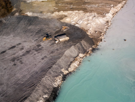 A cement factory tailings site with heavy machinery operating near a turquoise lake. Trucks and bulldozers move materials across the barren, gray terrain, highlighting industrial processes and environmental impactの写真素材