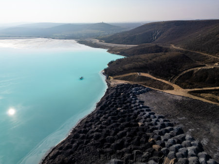 A turquoise tailings pond near a cement factory, surrounded by rugged hills. The vivid water contrasts with the barren land, creating a striking yet industrial scene of environmental impactの写真素材