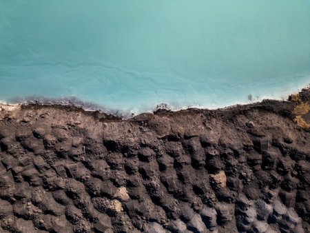 A striking contrast between turquoise water and dark, rugged terrain at a tailings lake. The industrial site reveals layers of environmental impact, blending sharp textures with vivid colorsの写真素材