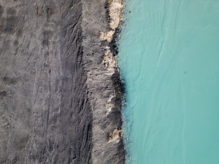A striking contrast between turquoise water and dark, rugged terrain at a tailings lake. The industrial site reveals layers of environmental impact, blending sharp textures with vivid colorsの写真素材