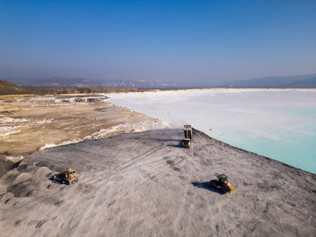 A cement factory tailings site with heavy machinery operating near a lake. Trucks and bulldozers move materials across the barren, gray terrain, highlighting industrial processes and environmental impactの写真素材