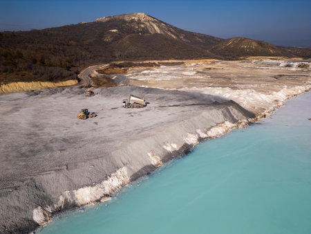 A cement factory tailings site with heavy machinery operating near a turquoise lake. Trucks and bulldozers move materials across the barren, gray terrain, highlighting industrial processes and environmental impactの写真素材