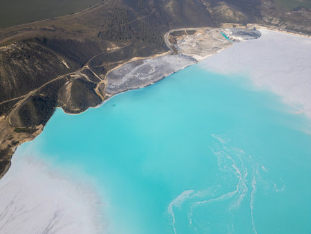 A turquoise tailings pond near a cement factory, surrounded by rugged hills. The vivid water contrasts with the barren land, creating a striking yet industrial scene of environmental impactの写真素材