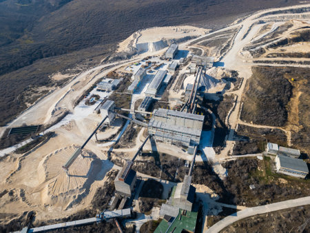 A large industrial cement plant surrounded by barren hills and dirt roads. Conveyor belts and storage facilities dominate the site, showcasing active production and its environmental impactの写真素材
