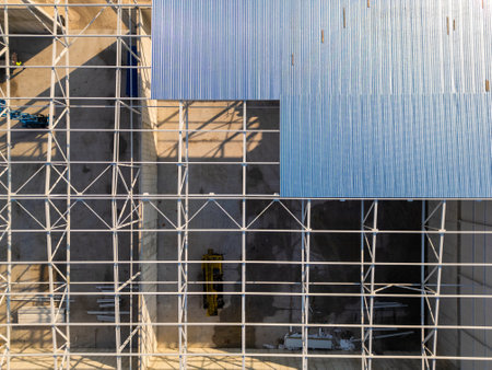 A construction site with a partially built warehouse featuring a steel framework and metal roof. Workers and heavy machinery operate on-site, showcasing industrial progress and structural developmentの写真素材