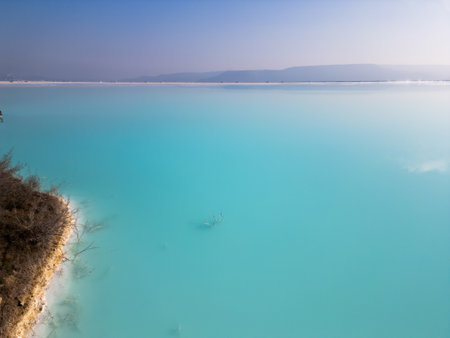 A turquoise pond near a cement factory, surrounded by rugged hills. The vivid water contrasts with the barren land, creating a striking yet industrial scene of environmental impactの写真素材