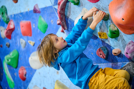 A boy in a blue hoodie climbs an indoor bouldering wall, gripping colorful holds with focus and determination. He reaches for the next hold, showcasing perseverance, strength, and adventureの写真素材