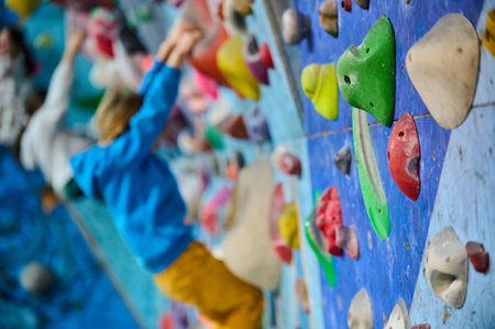 Indoor climbing gym with a focus on colorful holds. In the blurred background, a young boy in a blue hoodie and yellow pants is climbing, adding depth and action to the sceneの写真素材