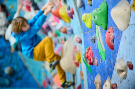 Indoor climbing gym with a focus on colorful holds. In the blurred background, a young boy in a blue hoodie and yellow pants is climbing, adding depth and action to the sceneの写真素材