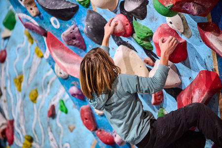 A young boy in a green hoodie climbs an indoor bouldering wall, gripping colorful holds. He focuses on his next move, demonstrating determination, strength, and an adventurous spirit in a climbing gymの写真素材