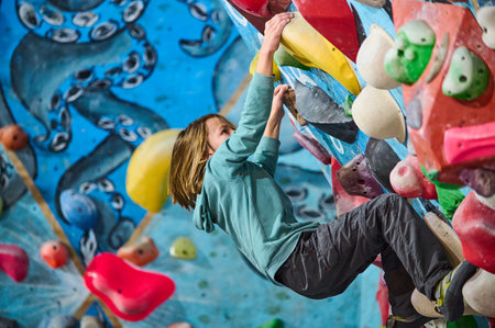 A young boy in a green hoodie climbs an indoor bouldering wall, gripping colorful holds. He focuses on his next move, demonstrating determination, strength, and an adventurous spirit in a climbing gymの写真素材