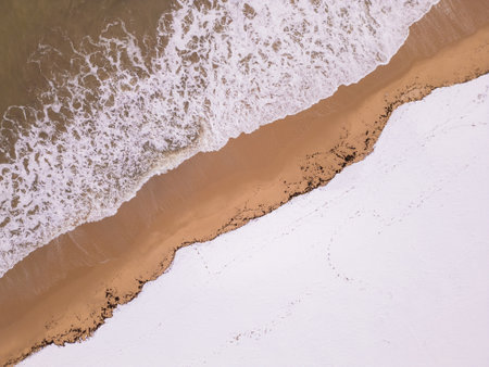 An overhead perspective of a winter shoreline where foamy ocean waves meet snowy land. The combination of textures and colors highlights the beauty of seasonal transitions in nature.の写真素材