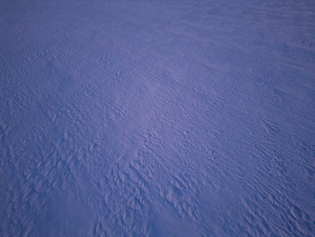 Aerial view of a textured snow-covered surface shaped by wind patterns. The frozen landscape showcases intricate natural formations, creating a serene and abstract winter scene under soft atmospheric lightの写真素材