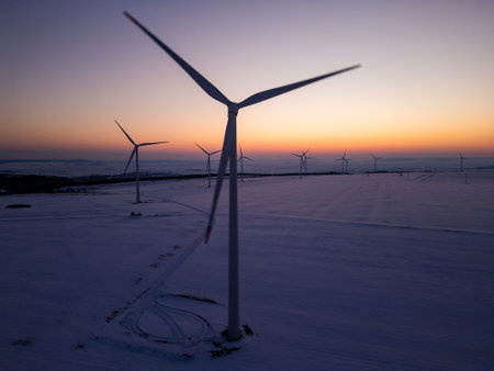 Aerial view of a wind farm in a snow-covered landscape at sunset. Wind turbines stand tall against the warm glow of the setting sun, creating a contrast between renewable energy and the winter sceneryの写真素材