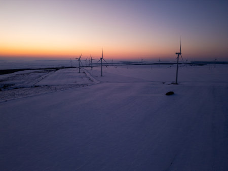 Aerial view of a wind farm in a snow-covered landscape at sunset. Wind turbines stand tall against the warm glow of the setting sun, creating a contrast between renewable energy and the winter sceneryの写真素材