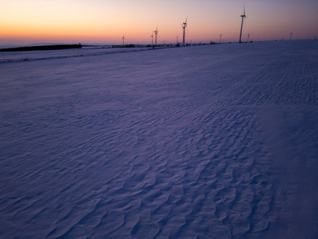 Aerial view of a wind farm in a snow-covered landscape at sunset. Wind turbines stand tall against the warm glow of the setting sun, creating a contrast between renewable energy and the winter sceneryの写真素材