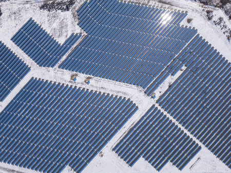 Aerial view of a large solar farm covered in snow, surrounded by a rural landscape with scattered houses and trees. The solar panels contrast against the white ground, showcasing renewable energy in winter conditionsの写真素材