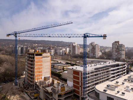 Construction site with a high-rise building under development and two large tower cranes. Urban landscape with residential buildings, roads, and greenery in the background, showing city growth and infrastructureの写真素材