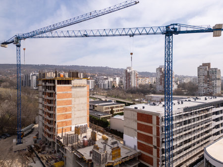 Construction site with a high-rise building under development and two large tower cranes. Urban landscape with residential buildings, roads, and greenery in the background, showing city growth and infrastructureの写真素材