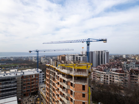 Construction site with a high-rise building under development and two large tower cranes. Urban landscape with residential buildings, roads, and greenery in the background, showing city growth and infrastructureの写真素材