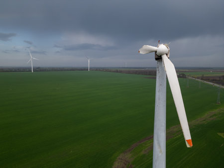 Close-up of a damaged wind turbine blade on a large green field with multiple turbines and cloudy sky in the background, symbolizing energy issues and maintenance needsの写真素材