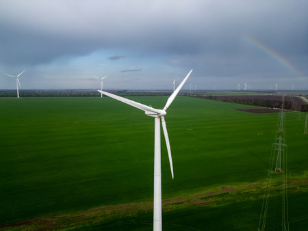 Close-up aerial view of a wind turbine against a backdrop of vast green fields and other turbines,...