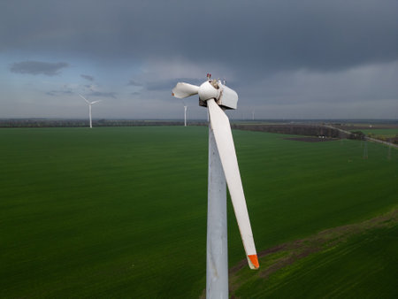 Close-up of a damaged wind turbine blade on a large green field with multiple turbines and cloudy sky in the background, symbolizing energy issues and maintenance needsの写真素材