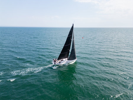Sailing yacht with black sails cruising on open sea with distant coastline under cloudy sky, aerial viewの写真素材