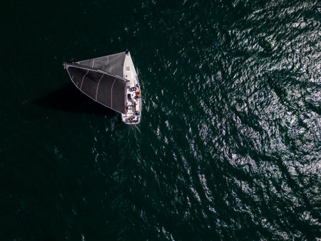 Top view of a sailing yacht with black sails moving through open sea, leaving a white wake behind on shimmering green waterの写真素材