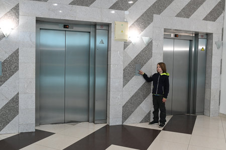 11 year old boy pressing an elevator button in a modern building lobby with stainless steel doors and geometric tile wallsの写真素材