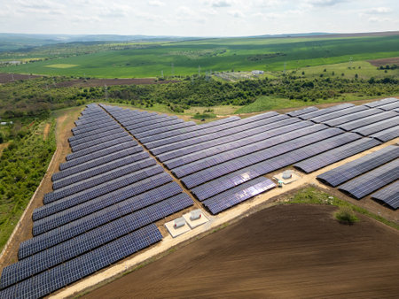 Aerial view of a large solar power farm with rows of photovoltaic panels set in a rural landscape, surrounded by green fields and power lines. Renewable energy conceptの写真素材