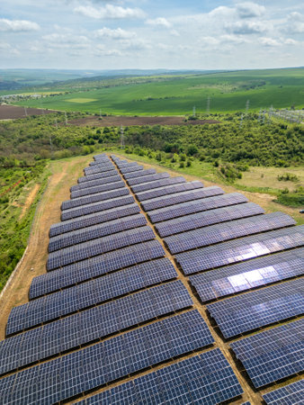 Aerial view of a large solar power farm with rows of photovoltaic panels set in a rural landscape, surrounded by green fields and power lines. Renewable energy conceptの写真素材