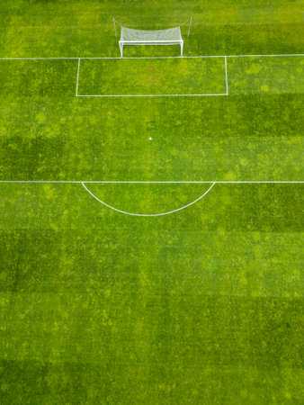 Aerial view of a green football field with white markings and a single goalpost. The grass shows...