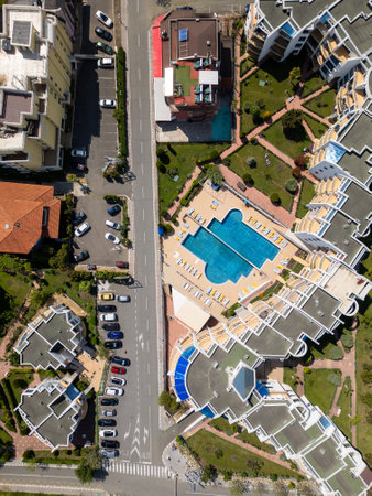 Aerial top-down view of a modern residential complex with a swimming pool, sun loungers, landscaped gardens, and surrounding buildings. Sunny day, geometric layout and parked cars visibleの写真素材
