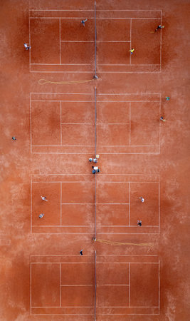 Aerial top-down view of multiple red clay tennis courts with players practicing. Symmetrical sports layout with lines, nets, and visible training activity on a sunny dayの写真素材