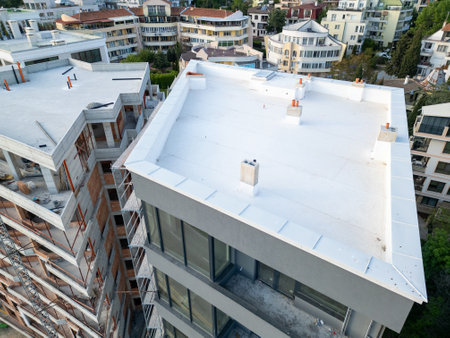 Aerial view of modern residential buildings under construction, featuring flat white rooftops, scaffolding, and unfinished facades. Surrounding neighborhood with completed homes in backgroundの写真素材