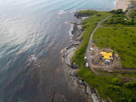 Aerial view of a coastal construction site on a green cliffside by the sea. Rugged shoreline, rock formations, and unfinished house with a yellow roof. Scenic and remote seaside landscapeの写真素材